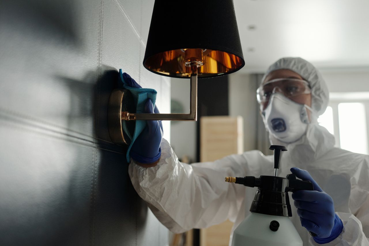 A person wearing protective gear sanitizes a wall lamp to prevent virus spread.